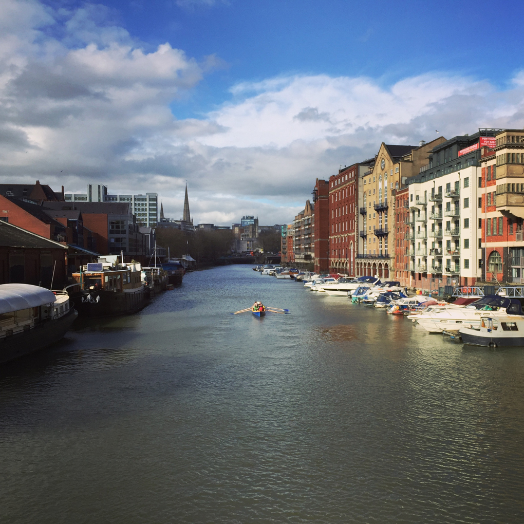 View of the river in Bristol, with buildings and boats on either side and a rowboat in the middle. Sunlight is hitting the water but there are clouds too.