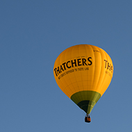 This is a photo of a hot air balloon with the Thatcher's cider branding on it. The balloon is yellow and green in a cloudless sky.