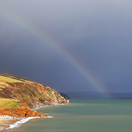 A faint rainbow emerging from a dark sea, curving inland over brightly-lit cliffs and coastal fields