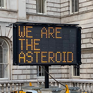 A dot-matrix roadworks sign displaying the slogan 'We are the asteroid'