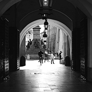 Black and white photo of a man cycling through some arches in the old market square in Krakow.