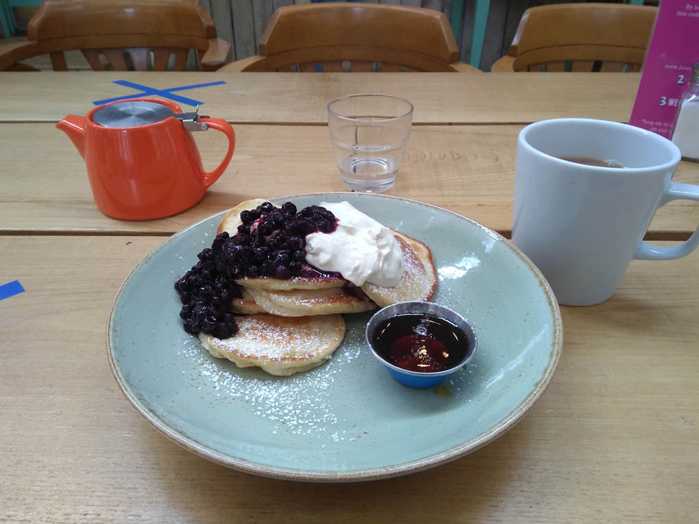 A plate of pancakes with blueberries, syrup and yogurt. Plus a pot of tea.