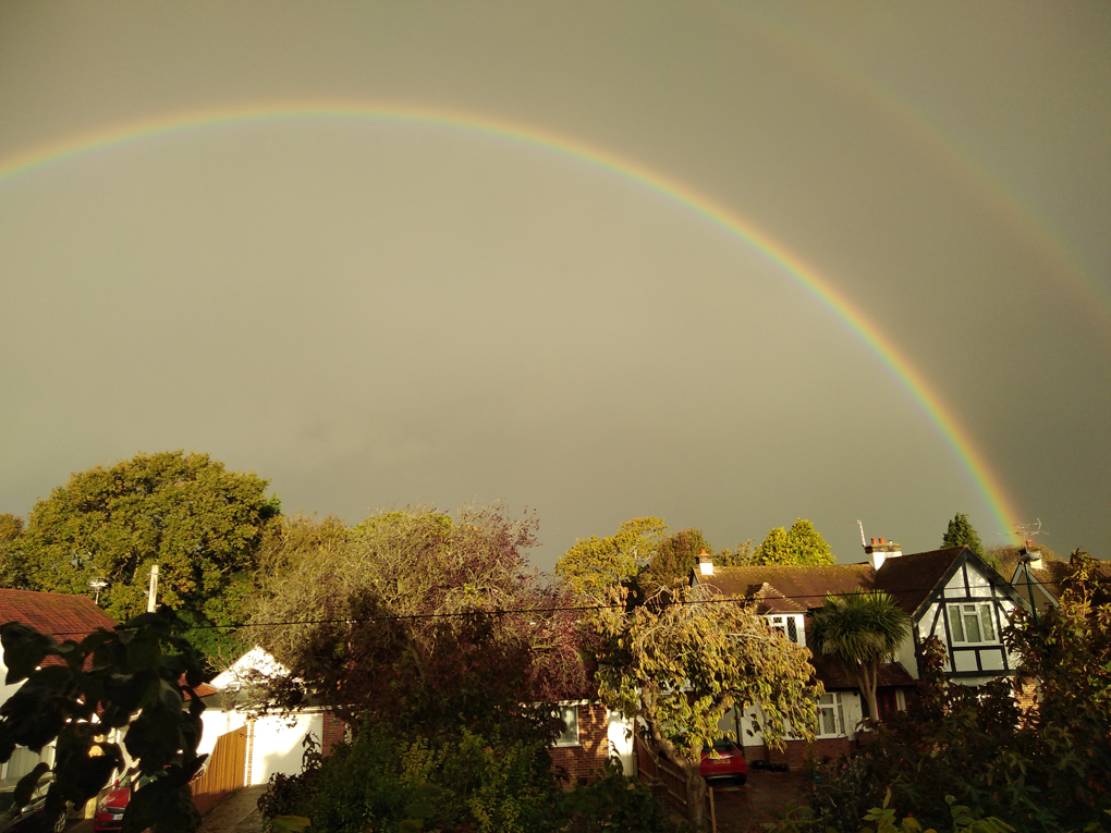 A rainbow arches over houses and trees against a background of a very dark sky