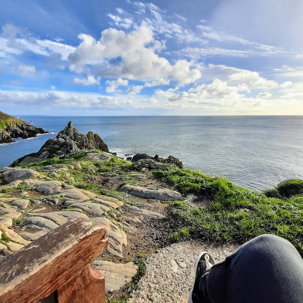 A view from a bench at the top of a dramatic cliff with jagged rocks leading to a bright calm sea and sky.