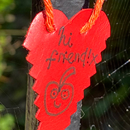 A paper heart has been tied to a metal gate. The text 'hi friend!' is written on the heart with a drawing of a smiling apple.