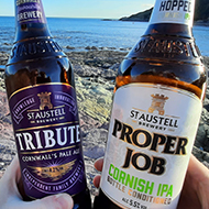 Two hands holding beer bottles of Tribute and Proper Job ale on a rocky beach with the sea in the background
