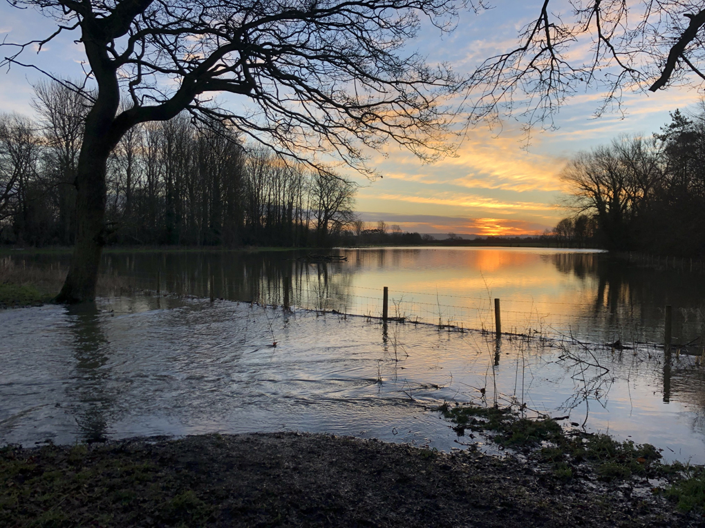 Sunrise over a flooded field, with water flowing from the field across a footpath in the foreground.