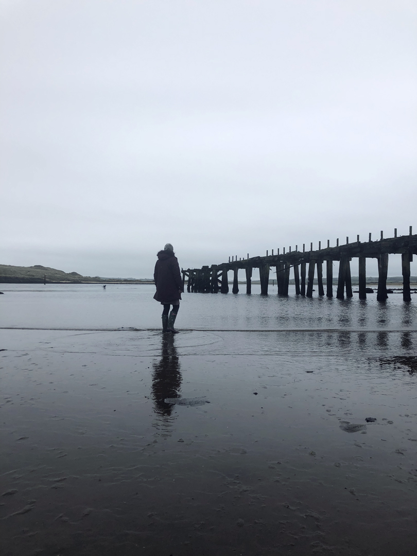A woman overlooking an estuary with rail bridge ruins silhouetted against the grey waters and skies.