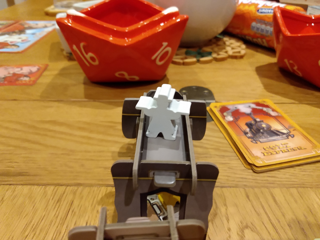 A white meeple (wooden person in a game) stands on the front of a cardboard train