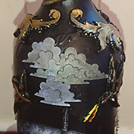 A wine bottle painted black and decorated Gothic style with a dark and daunting castle, made more eerie in the light of a bright moon complete with flying bats, dragons approaching the castle, dark clouds hanging over, and a snake entwined around the neck of the bottle. A dark rusty old gate features on the back, over which hangs an elaborate sign with the name Castle Skull on it. I used mainly air dried clay except for the clouds which are paper cut outs. Not bad for my first attempt, I think!