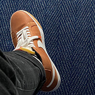 A first person perspective looking down at a blue hotel carpet. The person is sitting on a bed and you can only see their legs. They are wearing black jeans and orange trainers.