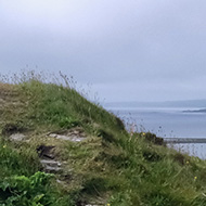 A small grassy cairn with an ancient stone entranceway, and a modern metal gate, stands high on a hillside overlooking the sea and small islands beyond.