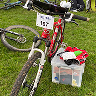 A mountain bike with a map board and attached race number (167) propped up against a box, in a field with other bikes.
