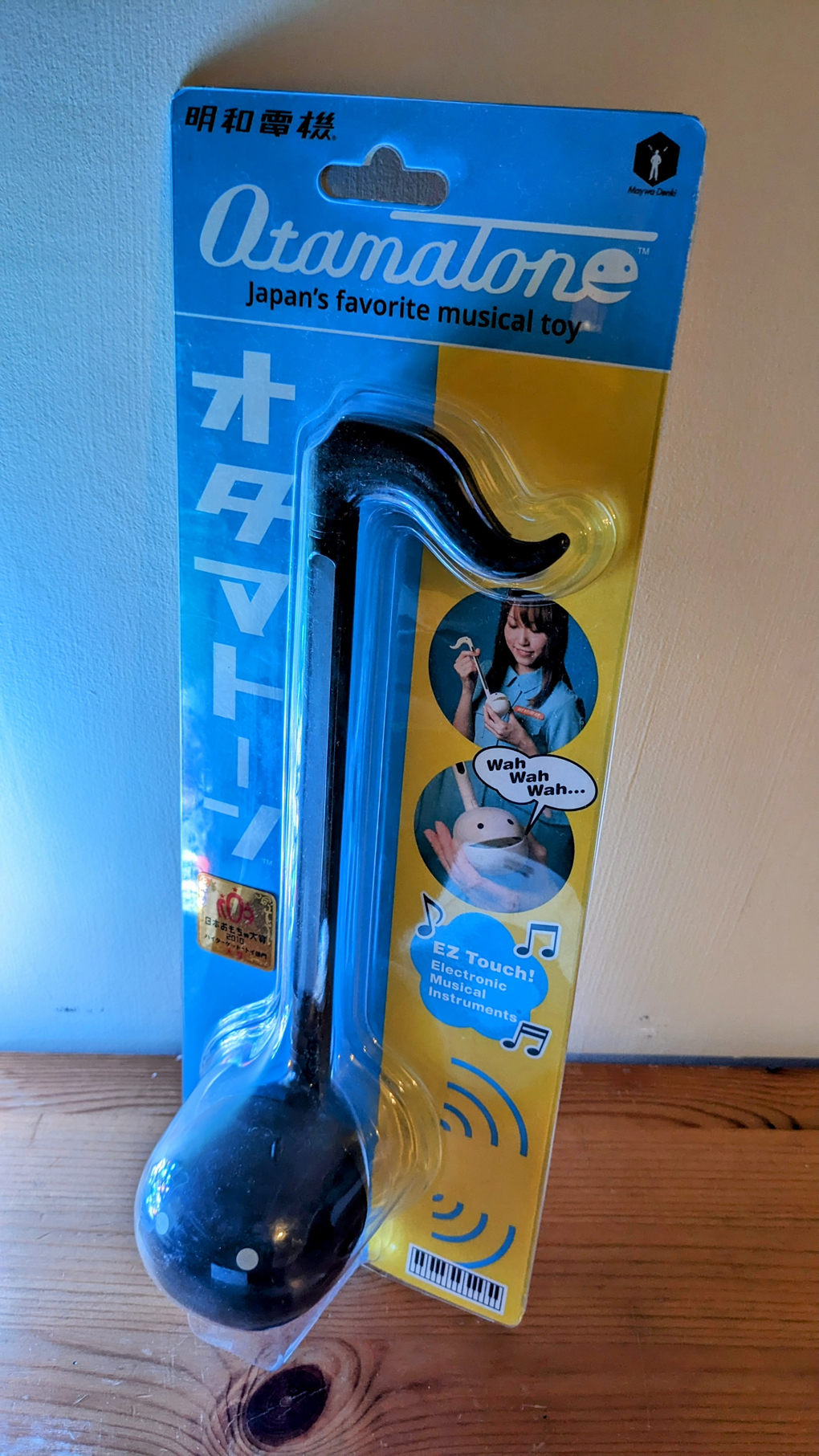 An Otamatone in it's packaging which demonstrates its use. It is shaped like a musical note. The stem is for choosing the note and the sound comes out of a little mouth/face drawn on the ball.