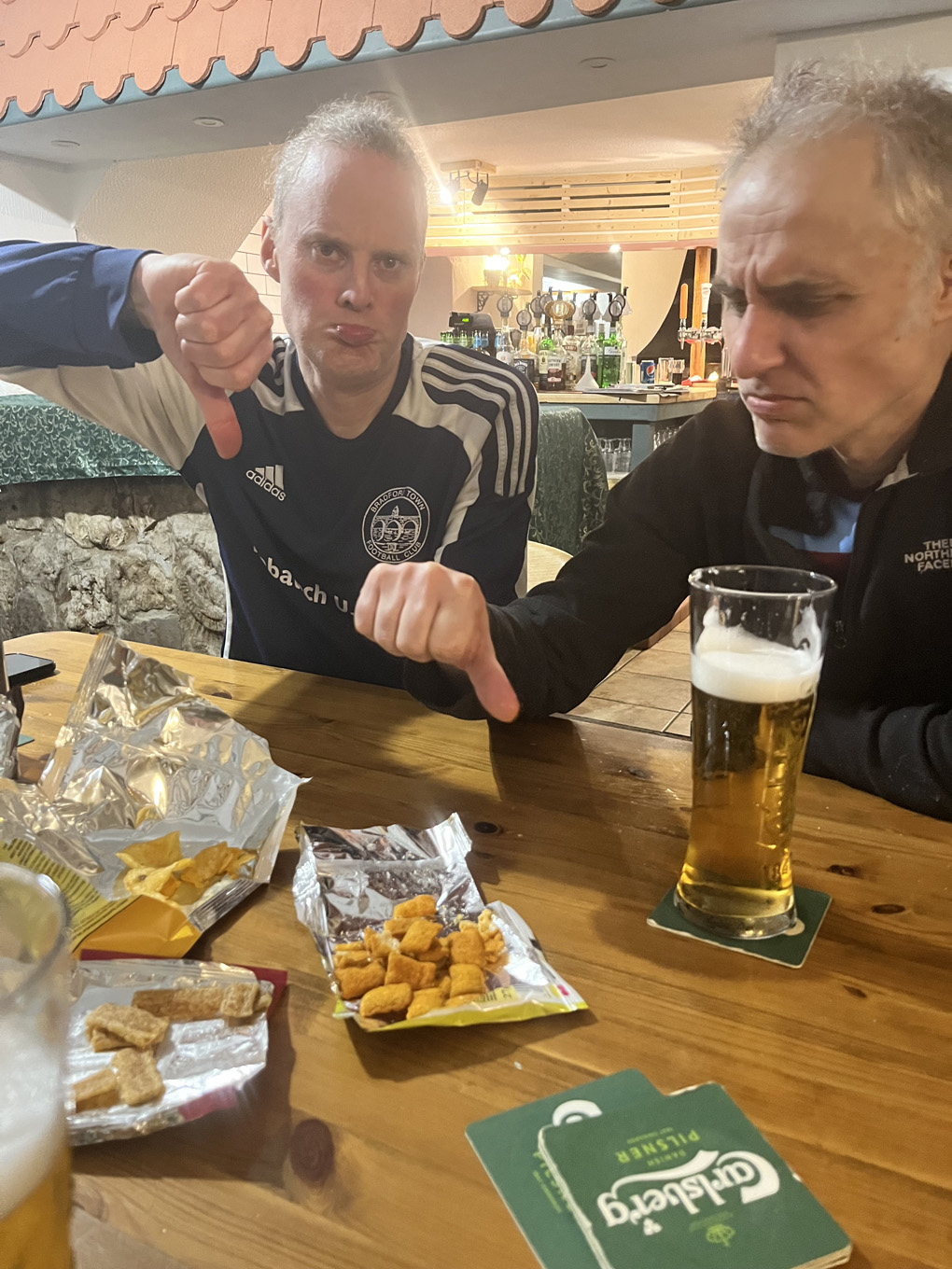 Two middle aged men in football shirts indicating their displeasure with the snacks on the table in front of them
