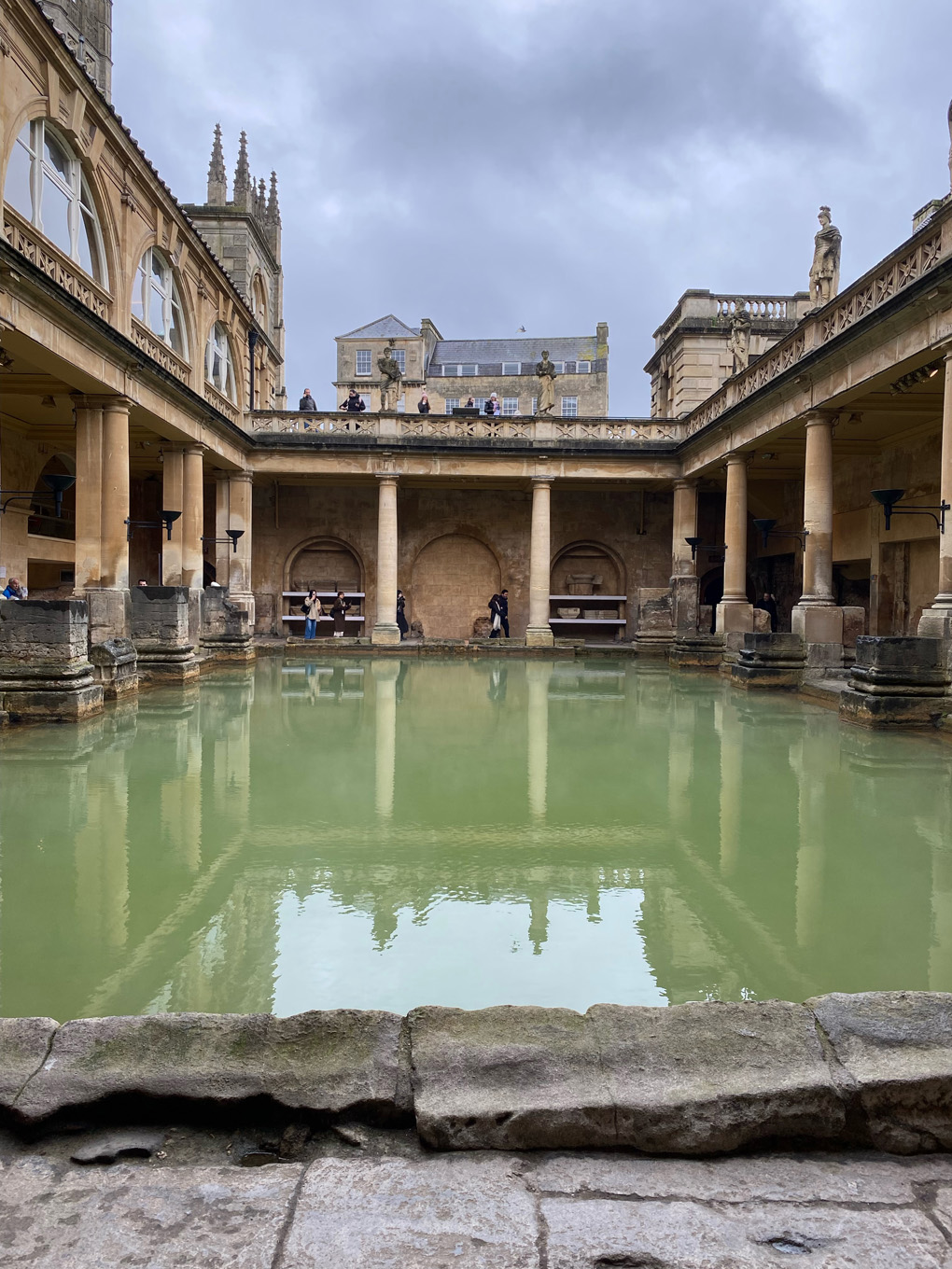 A view of the main bath at the Roman Baths in Bath. A rectangular pool of reflective greenish water is framed by sandstone collonades, pediments and statues of Romans. More sandstone buildings are looming in the background in front of a cloudy sky. A few tourists are standing under and above the collonades looking at the bath.