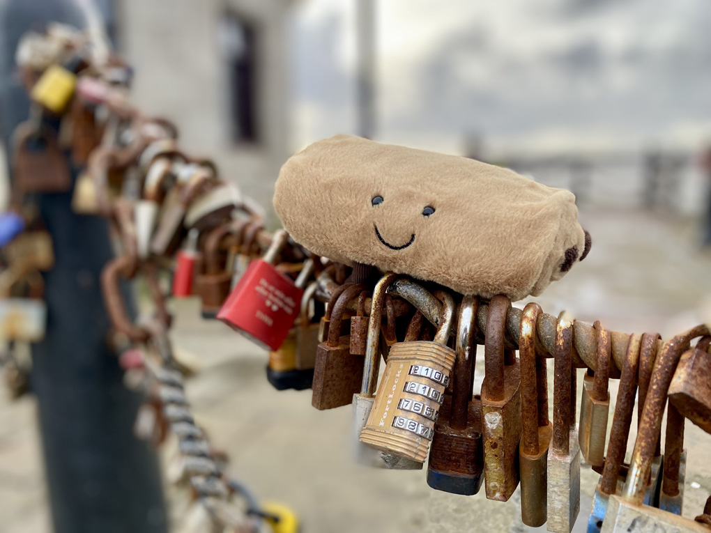 A soft toy pain au chocolat with a smiley face, sitting on a length of chain fence which is covered in padlocks.
