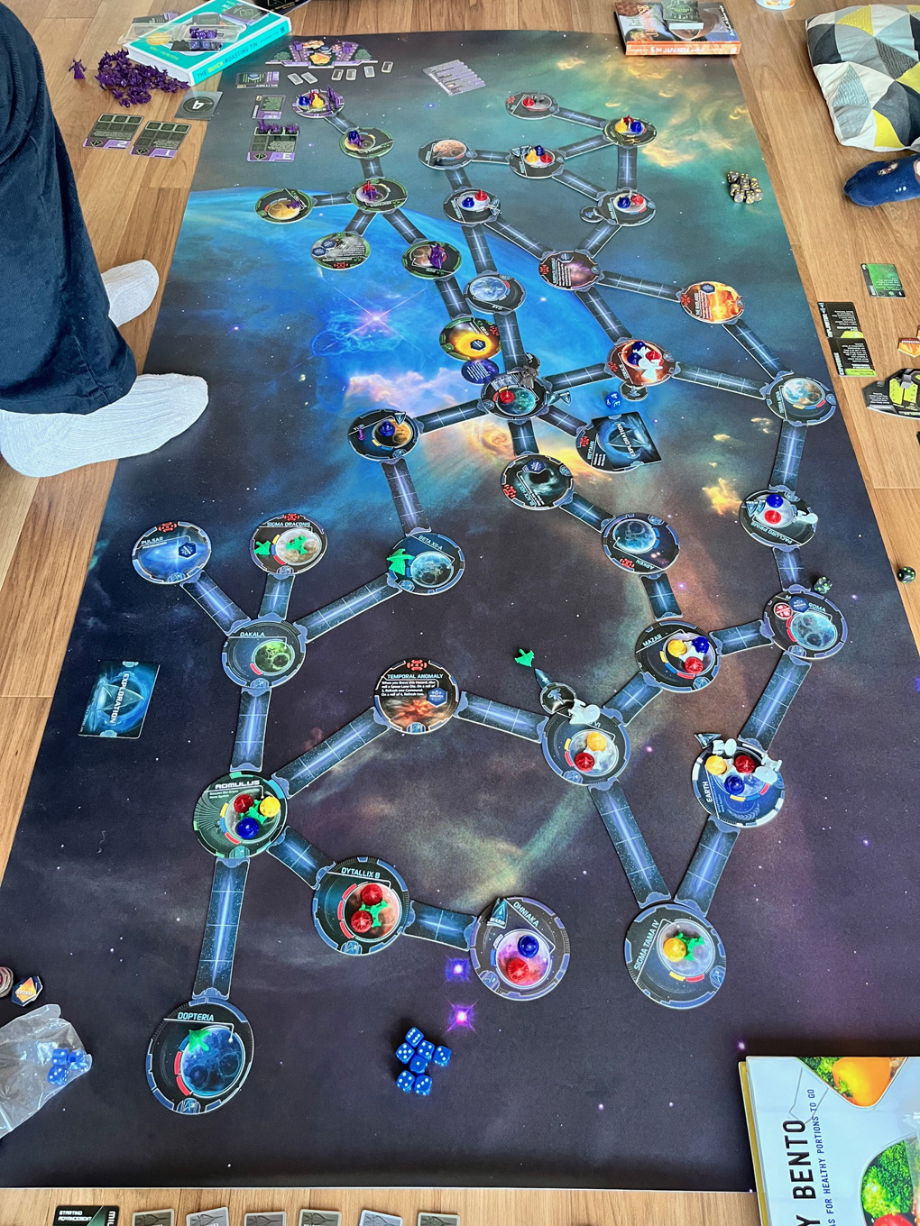 A large gameboard with galaxies, planets and other celestial bodies is spread over a wooden floor. The board is covered in counters, small model spaceships and other game parts.