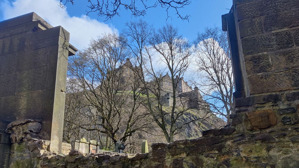 Looking up through a gap in an ancient wall stand bare trees in the early spring light and Edinburgh castle beyond against the blue sky