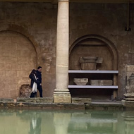 A view of the main bath at the Roman Baths in Bath. A rectangular pool of reflective greenish water is framed by sandstone collonades, pediments and statues of Romans. More sandstone buildings are looming in the background in front of a cloudy sky. A few tourists are standing under and above the collonades looking at the bath.
