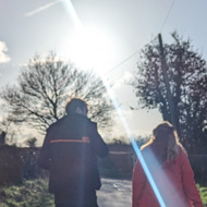 Three figures walking down a road with wide tall hedges on either side. The sun is in front of them leaving the figures almost as silhouettes