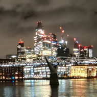 The city of London skyline at night is a forest of lights which reflect in the Thames by the Millennium footbridge.