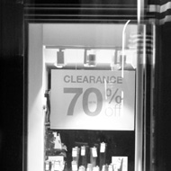 a homeless person sitting on the street in front of the bright window of a closed shop advertising a clearance sale