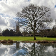 With a still lake in the foreground the sky, with streaks of white clouds, is reflected perfectly in the water. The composition is enhanced by the leafless tree on the far lake side, also reflected in the lake. In the distance the tower of the Cirencester Parish Church stands out.