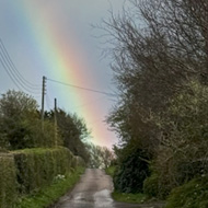 A brightly coloured rainbow appears at the end of a verdant country lane, with thick grey clouds covering the sky above