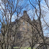 Looking up through a gap in an ancient wall stand bare trees in the early spring light and Edinburgh castle beyond against the blue sky