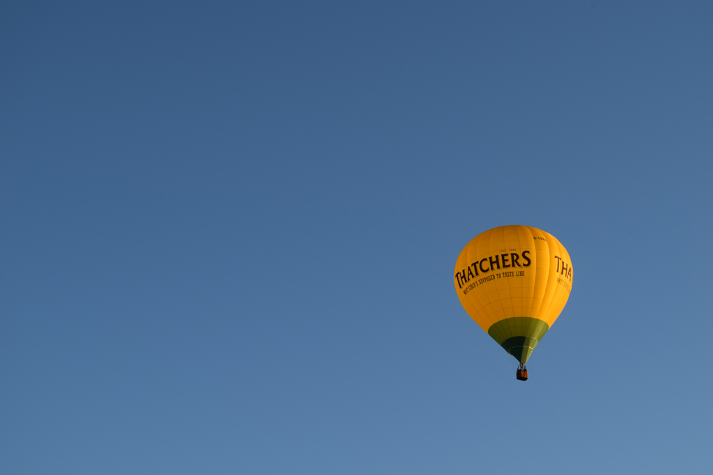 This is a photo of a hot air balloon with the Thatcher's cider branding on it. The balloon is yellow and green in a cloudless sky.