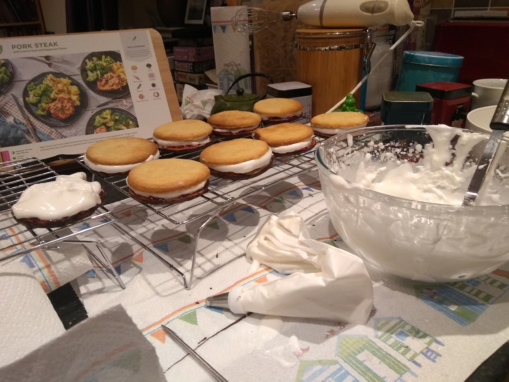 Cooling rack of uncoated wagon wheel biscuits on a cluttered kitchen worktop with bowl and icing bag containing marshmallow