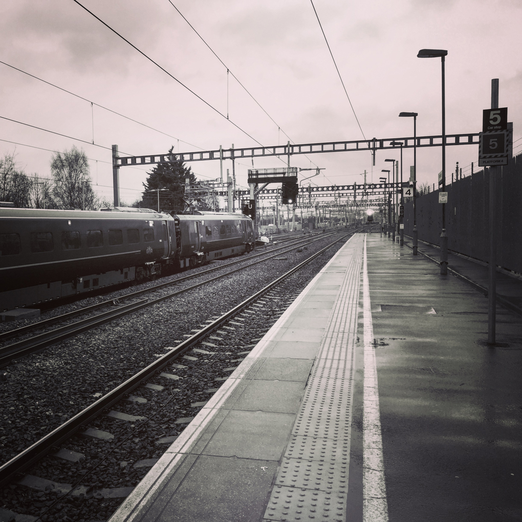 Looking down train tracks from the station platform, with aerial wires, tracks and markings converging in the distance