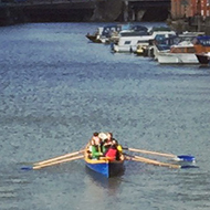 View of the river in Bristol, with buildings and boats on either side and a rowboat in the middle. Sunlight is hitting the water but there are clouds too.