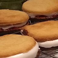 Cooling rack of uncoated wagon wheel biscuits on a cluttered kitchen worktop with bowl and icing bag containing marshmallow