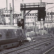 Looking down train tracks from the station platform, with aerial wires, tracks and markings converging in the distance