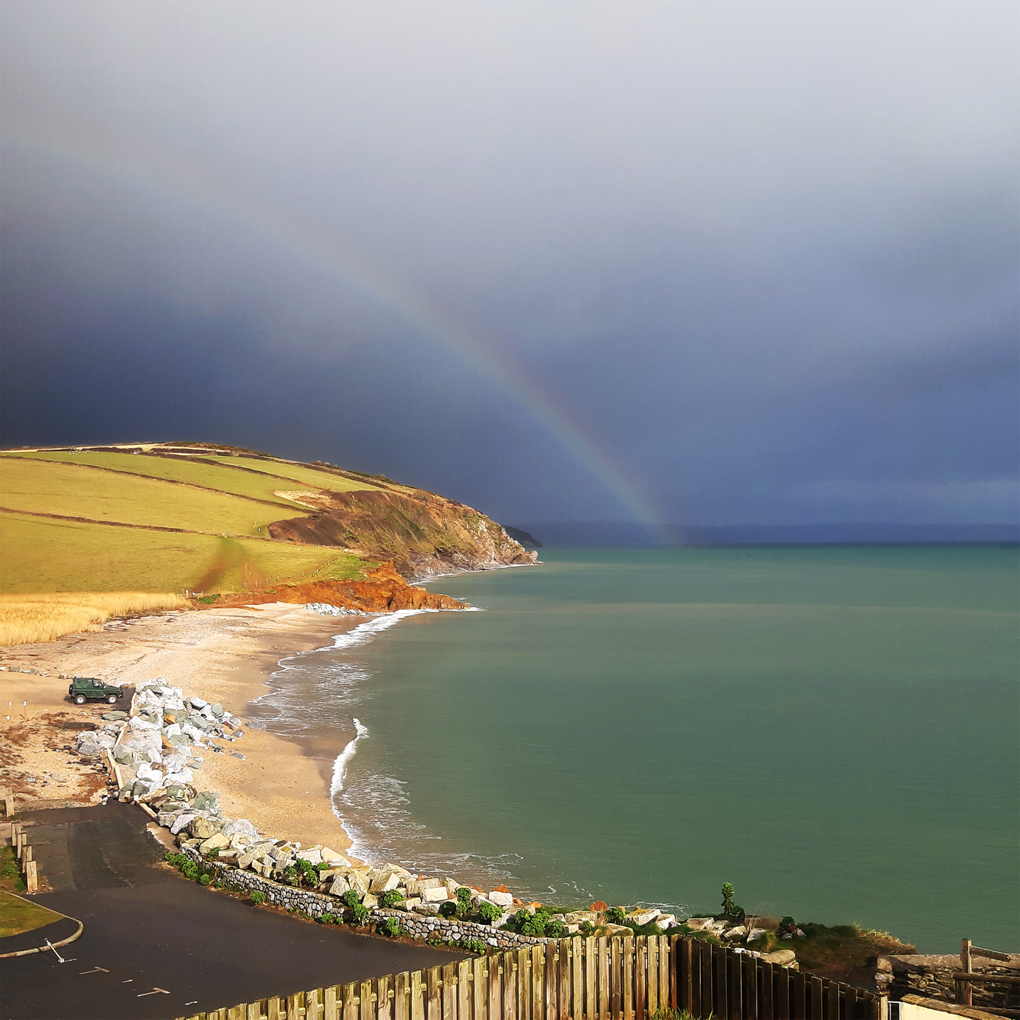 A faint rainbow emerging from a dark sea, curving inland over brightly-lit cliffs and coastal fields