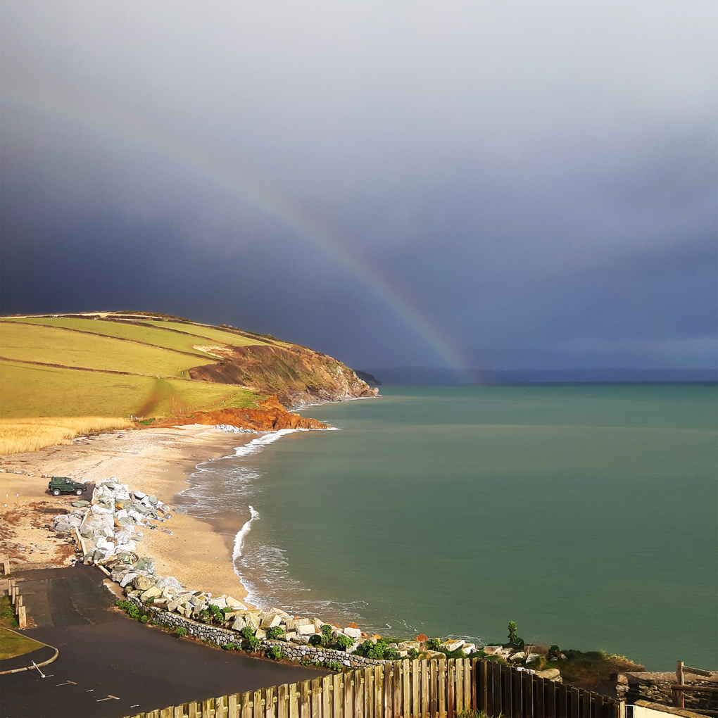 A faint rainbow emerging from a dark sea, curving inland over brightly-lit cliffs and coastal fields