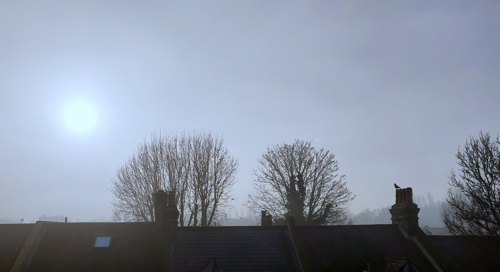 The sun behind misty cloud rises over terraced rooftops.