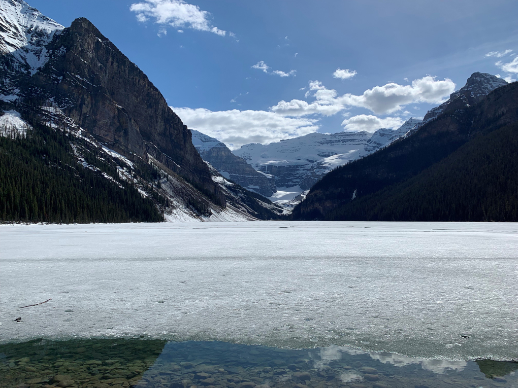 A frozen lake in the foreground with a snow capped mountain range behind