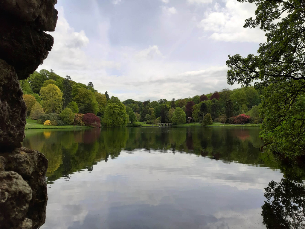 View across a still lake, with many different coloured trees surrounding an ornate bridge in the background, and a cloudy sky above