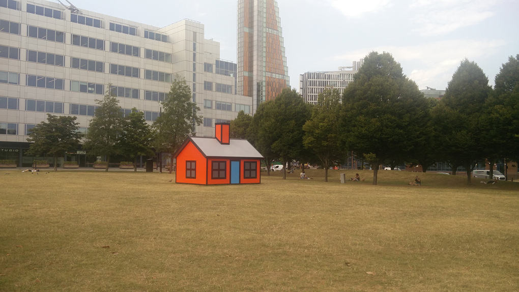 A 3-D model of a cartoon house on an otherwise empty field in a built-up area.