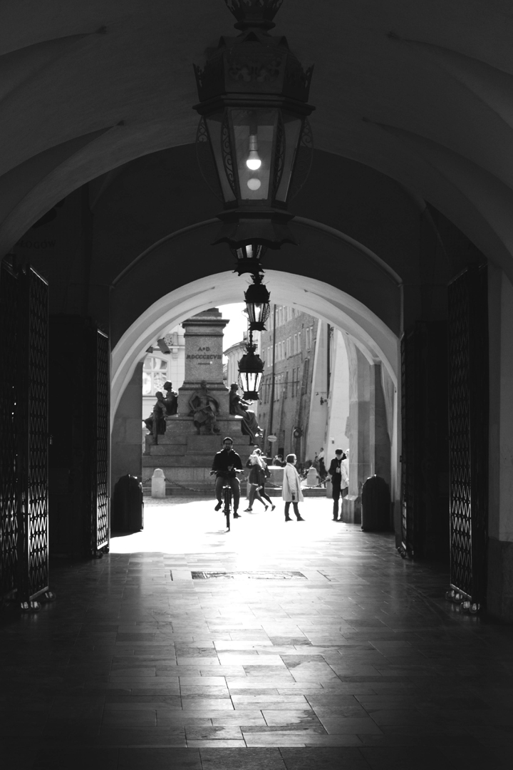 Black and white photo of a man cycling through some arches in the old market square in Krakow.