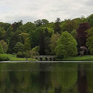 View across a still lake, with many different coloured trees surrounding an ornate bridge in the background, and a cloudy sky above