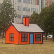 A 3-D model of a cartoon house on an otherwise empty field in a built-up area.