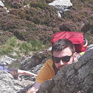 A shot of the mountains with a guy peeping round some rock he is climbing below.