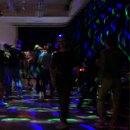 Lots of middle aged people jumping around in room lit with psychedelic lights