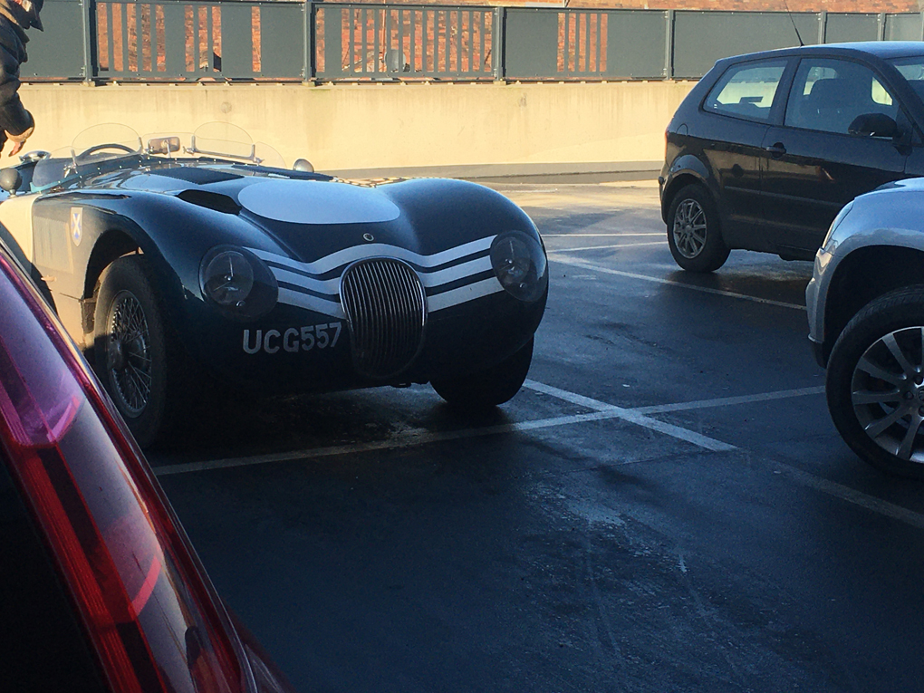 An open top classic Jaguar sports car is photographed between two modern vehicles in a supermarket car park.