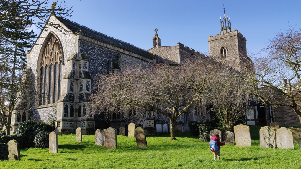 A small boy wandering in the sunny churchyard of a medieval flint and stone built church