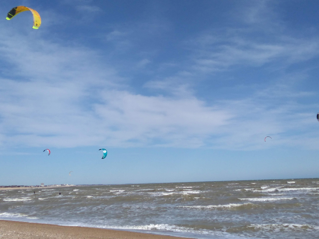It shows kite surfers on the shallow wavey beach with blue sky and wispy clouds above